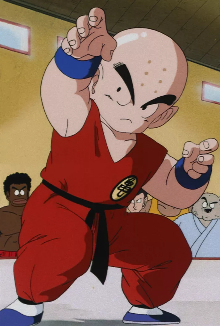 Portrait of a short, bald fighter with six forehead dots, wearing an orange gi and smiling nervously